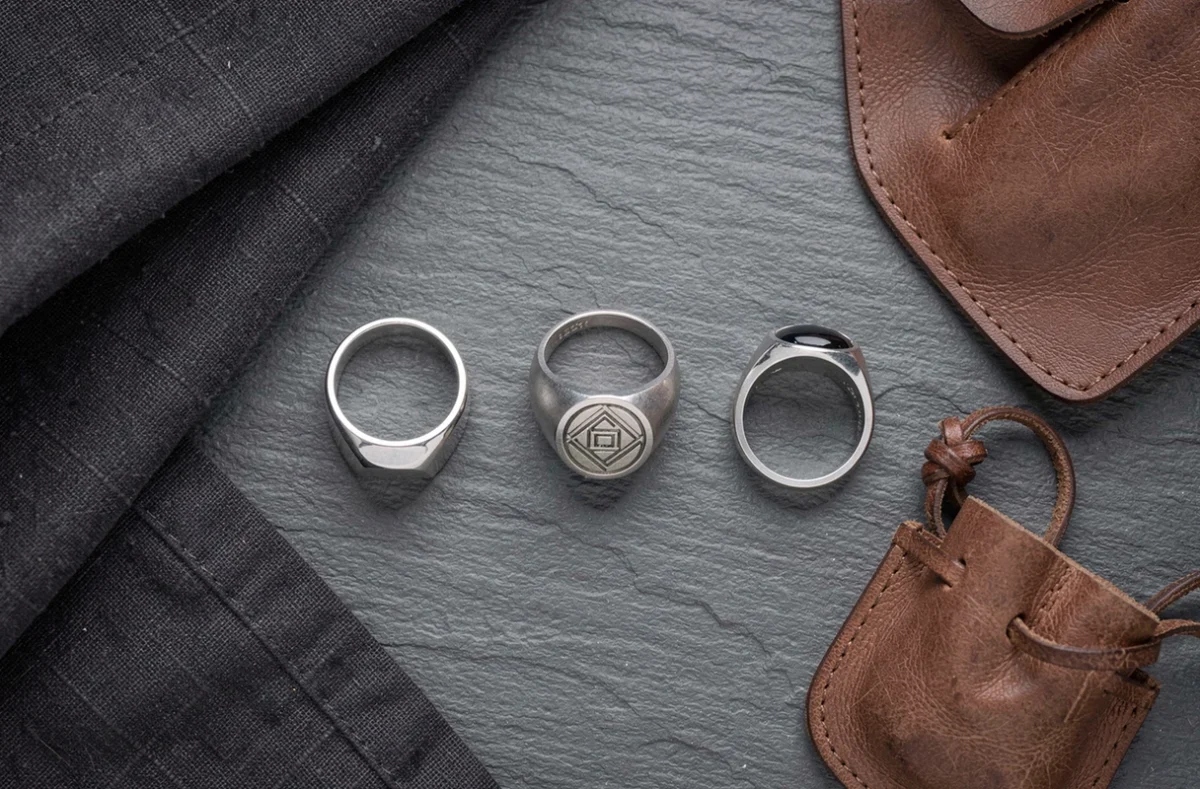 Flat lay de trois chevalières homme en argent aux designs variés sur ardoise grise avec accessoires en cuir et lin